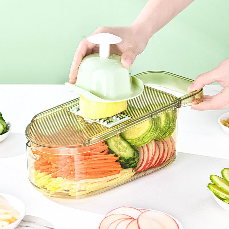 All-in-One Mandoline Slicer with clear container and green hand protector slicing yellow vegetable into thin strips over mixed sliced cucumbers, radishes, carrots in transparent base